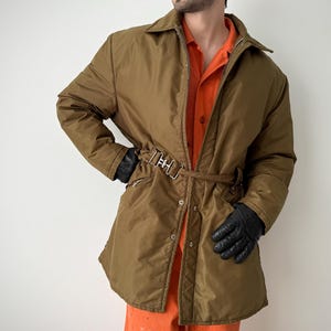 May include: A brown quilted coat with a belted waist and a collared neckline. The coat has a zip closure and is worn over an orange shirt. Black gloves complete the look.
