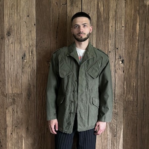 Puede incluir: Una chaqueta verde de estilo militar con cierre de botones y múltiples bolsillos. La chaqueta está hecha de un tejido de algodón duradero y tiene un corte holgado.