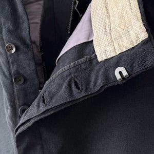 May include: Close-up of a dark navy blue garment, possibly a jacket or trousers, with visible buttons, buttonholes, and a metal clasp. The interior lining is a muted purple, and the collar features a textured off-white fabric.
