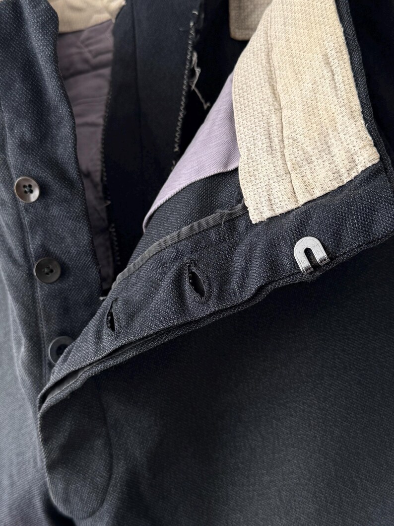 May include: Close-up of a dark navy blue garment, possibly a jacket or trousers, with visible buttons, buttonholes, and a metal clasp. The interior lining is a muted purple, and the collar features a textured off-white fabric.