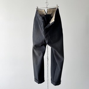 May include: Black trousers hanging from a metal hanger and chain against a white background. The trousers are buttoned at the waist and have cuffed hems. The interior lining is visible at the waist. The fabric appears to be a sturdy, dark material.