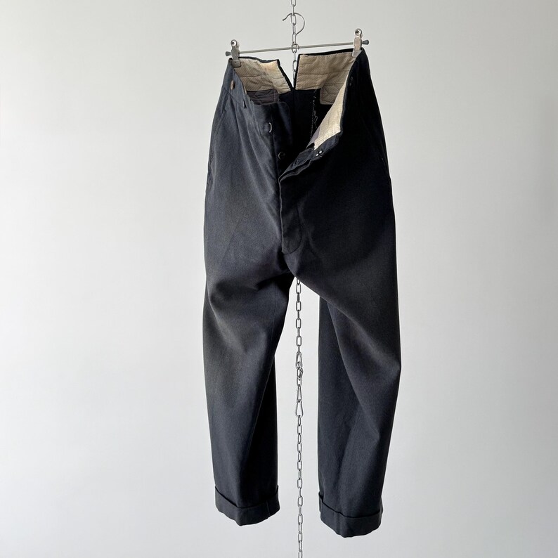 May include: Black trousers hanging from a metal hanger and chain against a white background. The trousers are buttoned at the waist and have cuffed hems. The interior lining is visible at the waist. The fabric appears to be a sturdy, dark material.