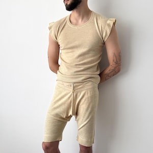 May include: A beige two-piece knitwear set. The top is a sleeveless shirt with frilled sleeves. The shorts have a button closure at the waist. The outfit is made of a soft, textured fabric.