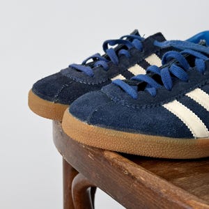 May include: A pair of navy blue suede sneakers with white stripes and blue laces. The sneakers have a brown rubber sole and are displayed on a wooden chair. The shoes are a classic athletic style.