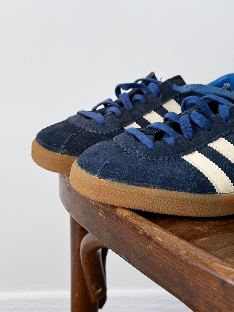 May include: A pair of navy blue suede sneakers with white stripes and blue laces. The sneakers have a brown rubber sole and are displayed on a wooden chair. The shoes are a classic athletic style.