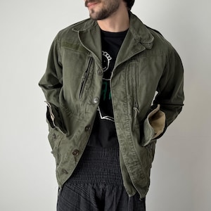 Puede incluir: Chaqueta verde oliva de estilo militar con múltiples bolsillos y cierres de botones. La chaqueta tiene cuello y un corte relajado. Las mangas están dobladas, revelando un forro beige. La persona lleva una camiseta negra y pantalones negros.