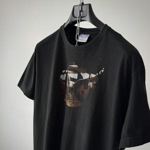 Puede incluir: Una camiseta negra de manga corta con un gráfico circular en el pecho. El gráfico contiene un paisaje en tonos marrón y blanco con la silueta de una persona y las letras FC. La camiseta se muestra colgada en una percha de plástico negra.
