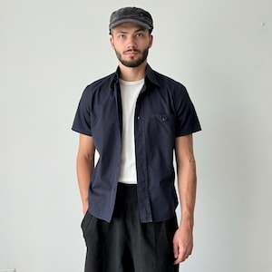Pode incluir: Um homem vestindo uma camisa azul escura de botões de manga curta com uma camiseta branca por baixo. Ele também está vestindo calças pretas e um boné preto.