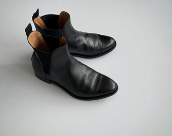 Może przedstawiać: Para czarnych skórzanych botków Chelsea. Buty mają elegancki, minimalistyczny design z elastycznymi panelami bocznymi i uchwytami. Podeszwy są czarne, a wnętrze ma jasnobrązowy kolor.