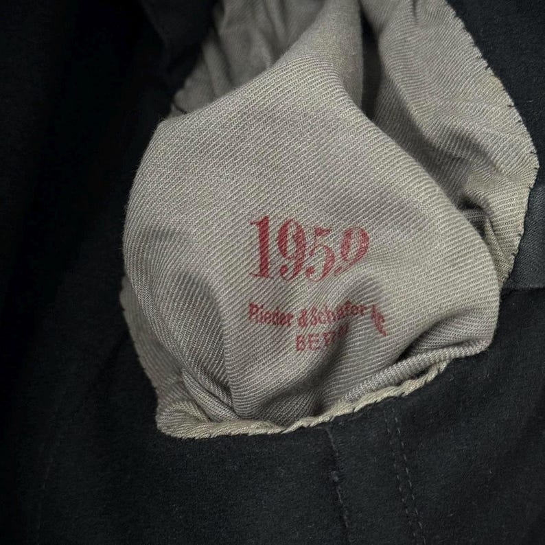 Puede incluir: Primer plano de una etiqueta de tela gris con el texto "1959" y "Rieder & Schaffer, Berl&iacute;n" en letras rojas.
