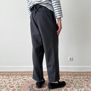 May include: Dark gray trousers with a high waist and button details. The pants have a relaxed fit and are cuffed at the ankles. The person is wearing black loafers and a striped long-sleeve shirt.