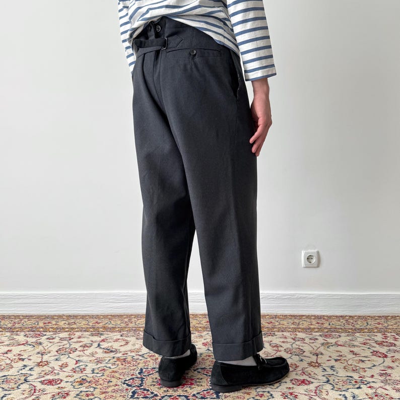 May include: Dark gray trousers with a high waist and button details. The pants have a relaxed fit and are cuffed at the ankles. The person is wearing black loafers and a striped long-sleeve shirt.