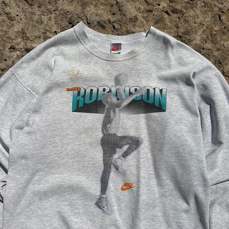 Puede incluir: Sudadera gris con un estampado gr&aacute;fico de un jugador de baloncesto en acci&oacute;n. El gr&aacute;fico presenta el texto "SWIFT ROBINSON" y un logotipo de Nike.