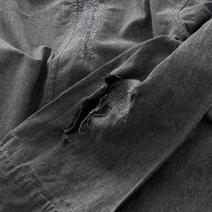 May include: Close-up of a distressed, charcoal-colored denim jacket. The jacket features visible wear and tear, with a large rip in the sleeve and distressed patches. The fabric appears soft and well-worn, suggesting a vintage or pre-loved item.