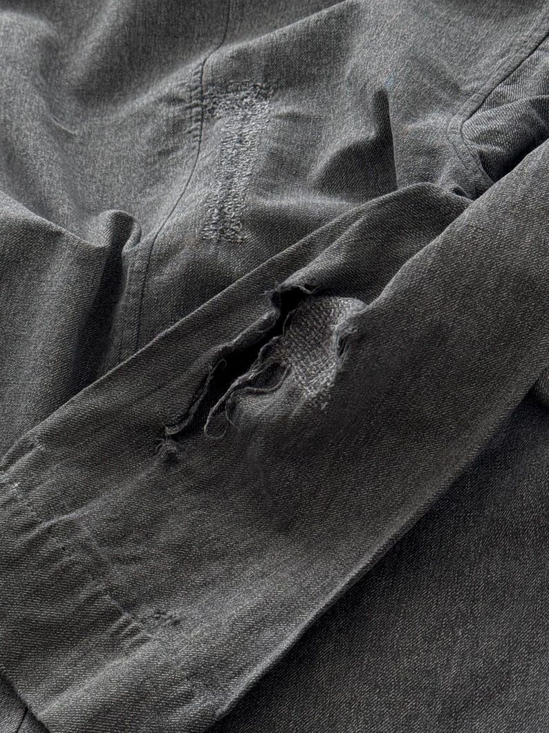 May include: Close-up of a distressed, charcoal-colored denim jacket. The jacket features visible wear and tear, with a large rip in the sleeve and distressed patches. The fabric appears soft and well-worn, suggesting a vintage or pre-loved item.