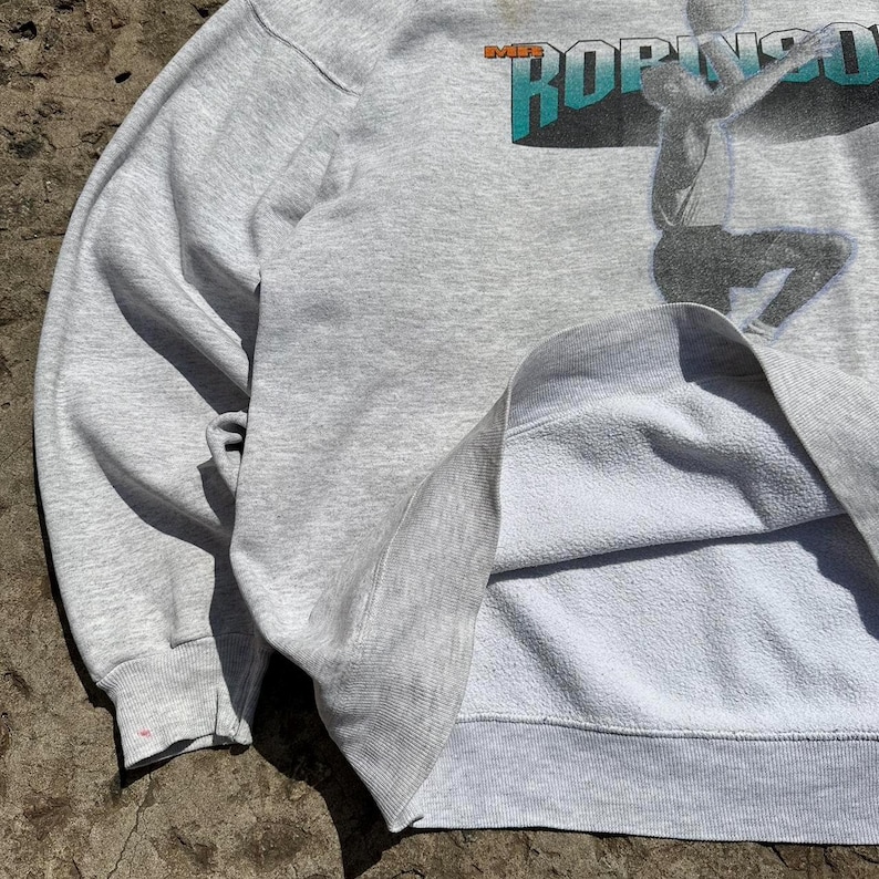 Puede incluir: Sudadera gris con un estampado gr&aacute;fico de un jugador de baloncesto con el texto "The Mike Robinson Show" en la parte delantera. La sudadera tiene un forro polar.