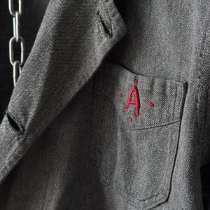 May include: Close-up of a gray denim jacket with a red embroidered "A" on the pocket. The jacket has a classic collar, button closure, and a silver chain detail. The fabric has a textured appearance.