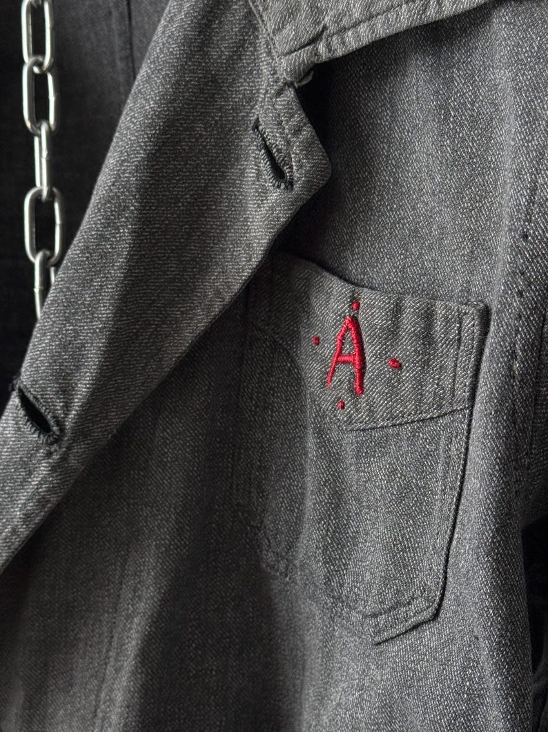 May include: Close-up of a gray denim jacket with a red embroidered "A" on the pocket. The jacket has a classic collar, button closure, and a silver chain detail. The fabric has a textured appearance.