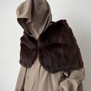 Puede incluir: Un abrigo con capucha de color beige con un cuello de piel marrón oscuro. El abrigo tiene un diseño sencillo con cierre de botón y una capucha. El cuello de piel añade un toque de lujo.