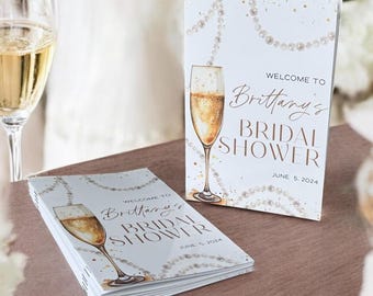 May include: A white bridal shower invitation with gold lettering and a pearl design. The invitation features a champagne flute with a gold rim and a glass of champagne. The text on the invitation reads "Welcome to Brittany's Bridal Shower June 5, 2024".