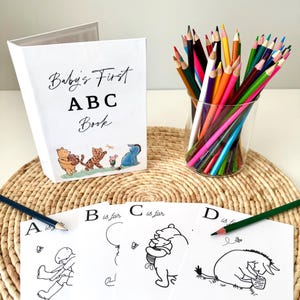 May include: A white "Baby's First ABC Book" with Winnie the Pooh characters and a glass of colored pencils. The book has the text "Baby's First ABC Book" on the cover. Coloring pages with letters and illustrations are also visible.