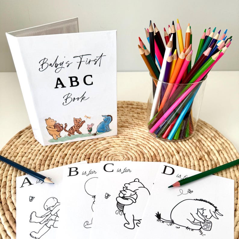 May include: A white "Baby's First ABC Book" with Winnie the Pooh characters and a glass of colored pencils. The book has the text "Baby's First ABC Book" on the cover. Coloring pages with letters and illustrations are also visible.