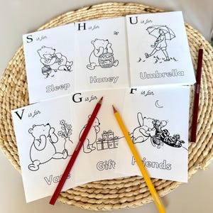 May include: A collection of white coloring pages featuring Winnie the Pooh characters and related words like "Sleep", "Honey", "Umbrella", "Vase", "Gift", and "Friends". Red and yellow colored pencils are placed on top of the pages.