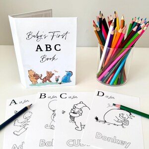 May include: A white "Baby's First ABC Book" with Winnie the Pooh characters, coloring pages, and a glass of colored pencils. The coloring pages feature letters and illustrations. A black pencil rests on the "A is for Ad" page.