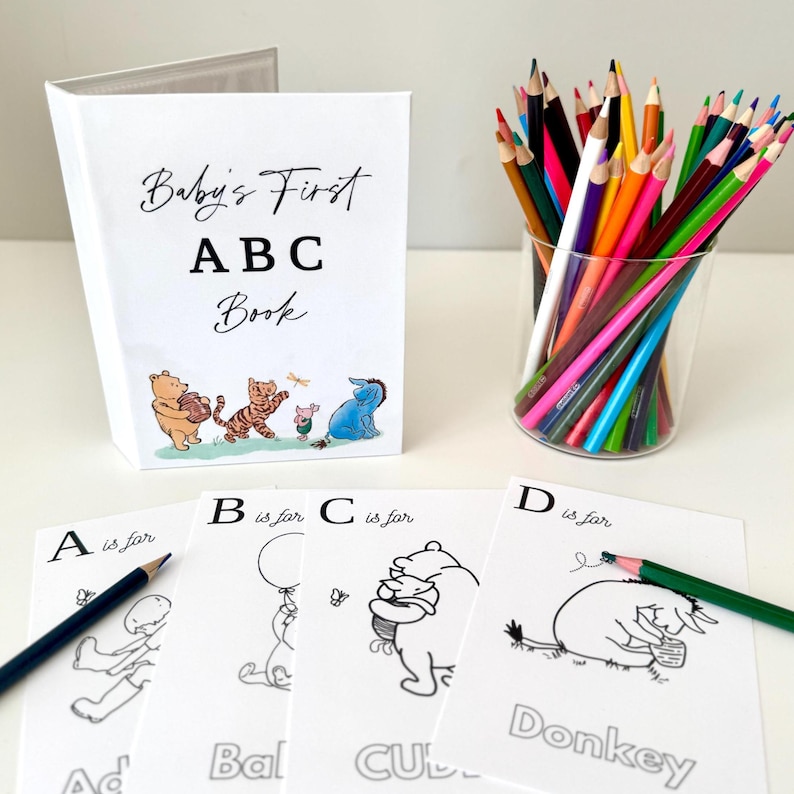 May include: A white "Baby's First ABC Book" with Winnie the Pooh characters, coloring pages, and a glass of colored pencils. The coloring pages feature letters and illustrations. A black pencil rests on the "A is for Ad" page.