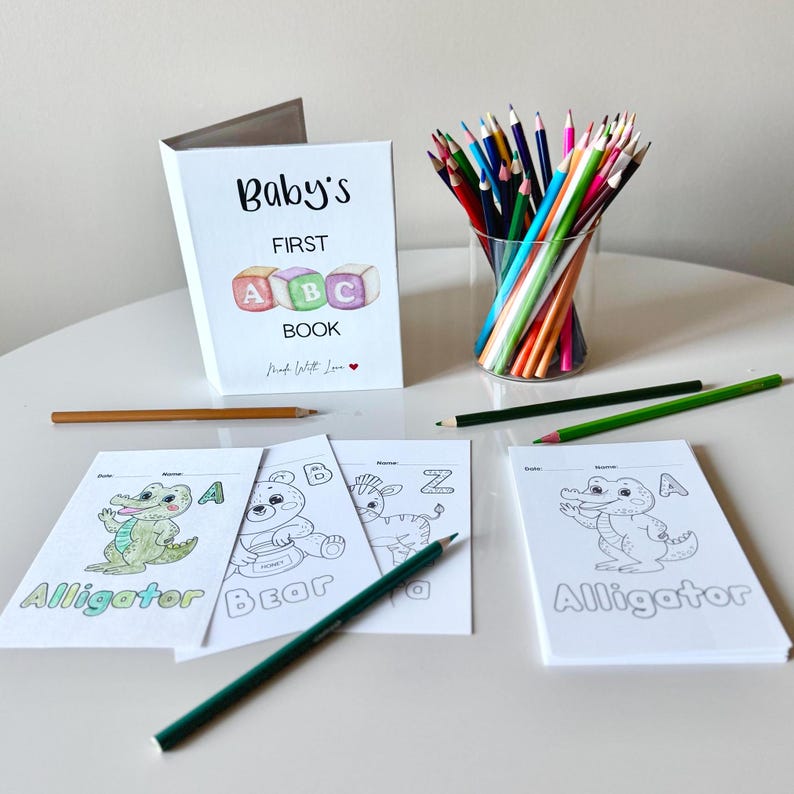 May include: A set of coloring pages featuring animals and letters. The pages are titled "Alligator", "Bear", and "Zebra". The pages are on a white table with a cup of colored pencils and a book titled "Baby's First ABC Book".