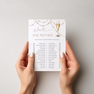 May include: A white card with gold text and a champagne glass illustration. The card reads "Would She Rather" and includes a list of choices for a bridal shower game. The card is held by two hands.