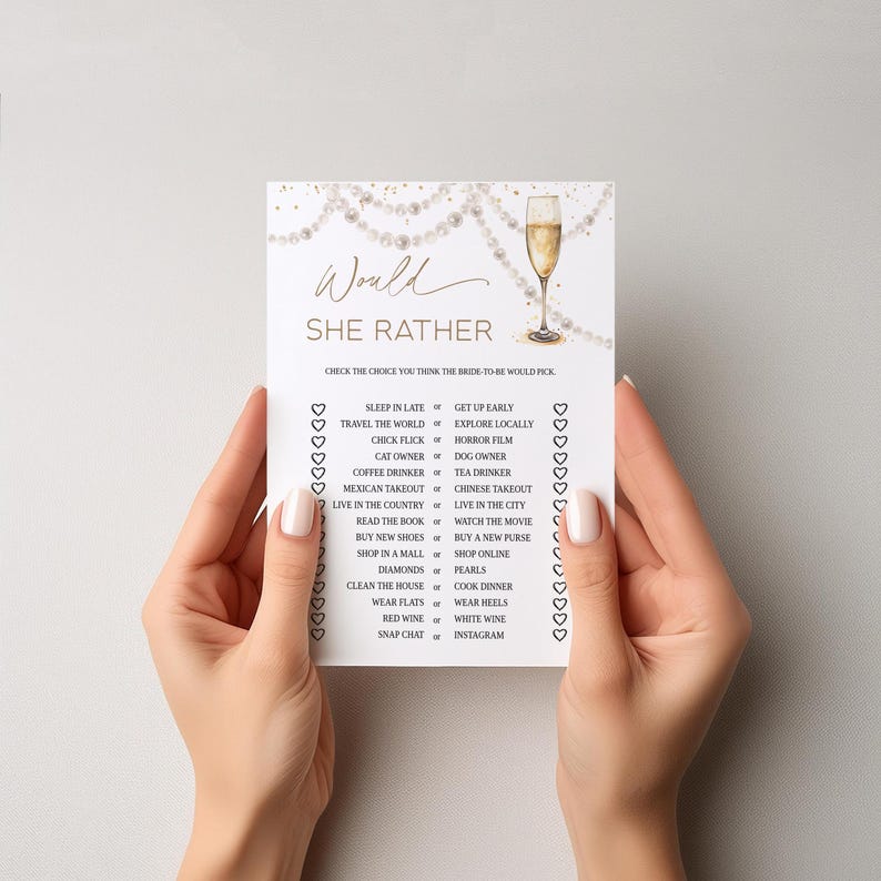 May include: A white card with gold text and a champagne glass illustration. The card reads "Would She Rather" and includes a list of choices for a bridal shower game. The card is held by two hands.