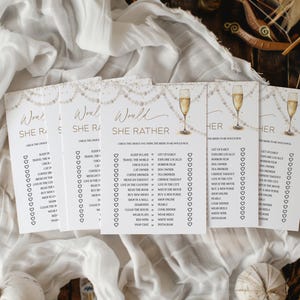 May include: A collection of white Would She Rather game cards with gold text and champagne glass illustrations. The cards are arranged on a white fabric background, perfect for a bridal shower or bachelorette party.