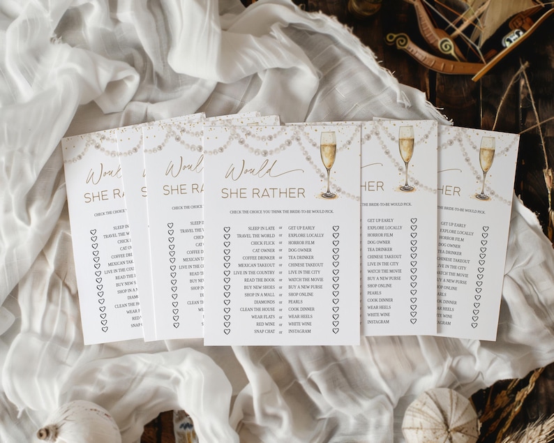 May include: A collection of white Would She Rather game cards with gold text and champagne glass illustrations. The cards are arranged on a white fabric background, perfect for a bridal shower or bachelorette party.