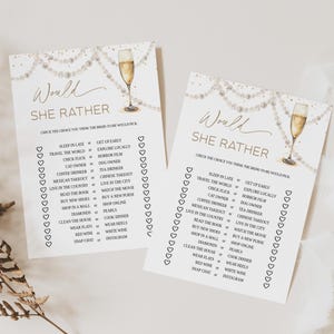 May include: Two white paper game cards with gold text and champagne glass illustrations. The cards read "Would She Rather" and list choices like "Sleep in Late or Get Up Early". Pearl string and heart accents.