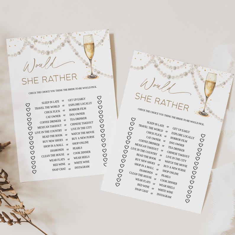 May include: Two white paper game cards with gold text and champagne glass illustrations. The cards read "Would She Rather" and list choices like "Sleep in Late or Get Up Early". Pearl string and heart accents.