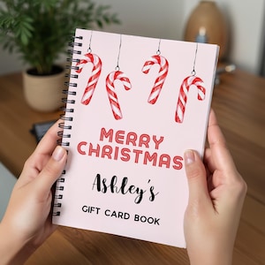 May include: A spiral-bound gift card book with a light pink cover. The cover features red and white candy canes hanging from the top, with the words "MERRY CHRISTMAS" in red and "Ashley's GIFT CARD BOOK" in black.