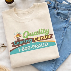 May include: Cream-colored sweatshirt with the text "Quality Learning Center 1-800-FRAUD" in green, brown, and blue. The design includes a treehouse graphic. Light blue jeans are folded next to the sweatshirt.