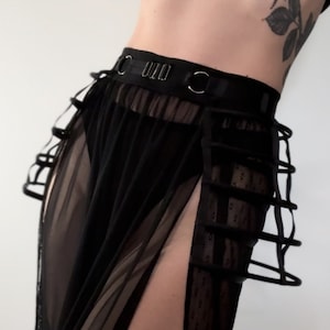 May include: Black sheer skirt with a high waist and cage-like detailing. The skirt features a slit on the side, revealing a glimpse of the leg. The skirt is made of a lightweight, see-through fabric.
