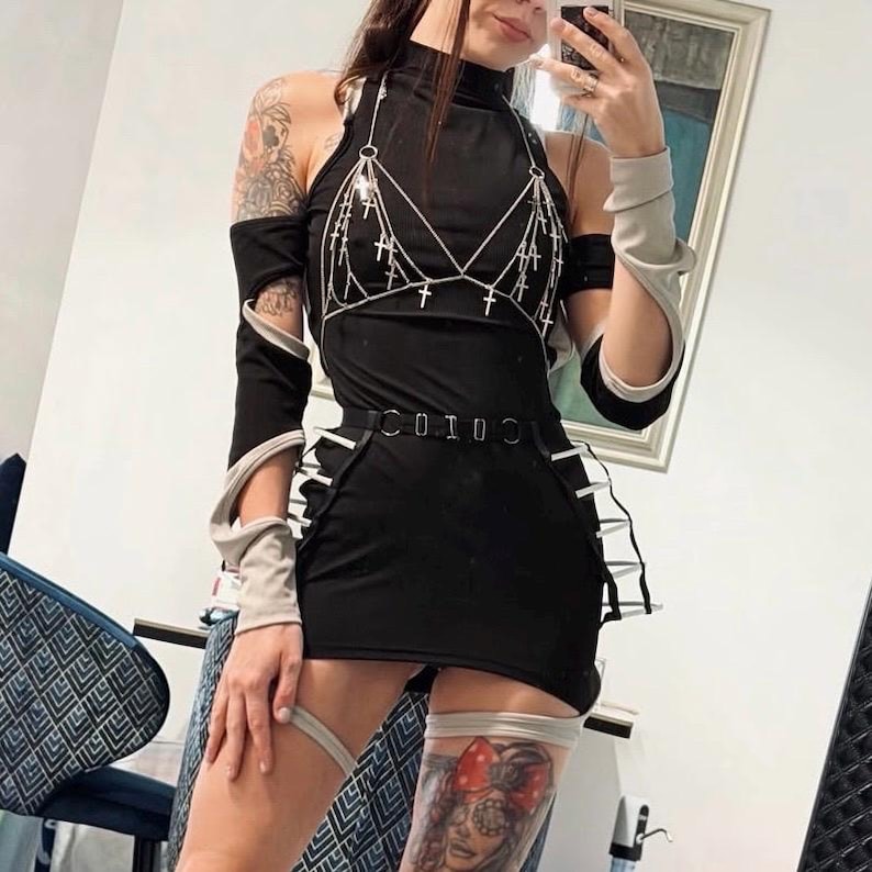 May include: Black mini dress with a high neckline and a silver chain harness adorned with cross pendants. The dress features a belt and white accents. The outfit includes matching arm warmers and leg garters.