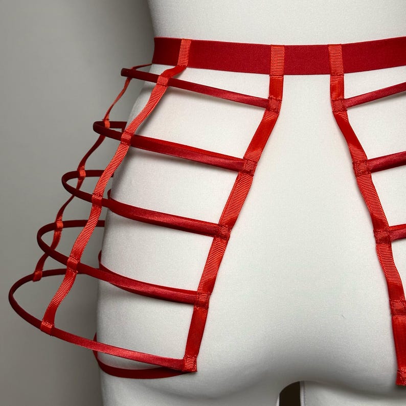 May include: A red lingerie harness featuring a cage-like design. The harness is made of red satin ribbon and elastic, with a wide waistband and vertical and horizontal straps creating a geometric pattern. The garment is displayed on a white mannequin.