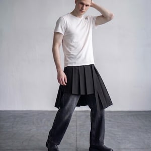 May include: A white short-sleeved crew neck t-shirt paired with a black pleated skirt worn over dark gray denim jeans. The skirt features sharp vertical pleats and a long, narrow fabric strip hanging down from the waistline on the right side.