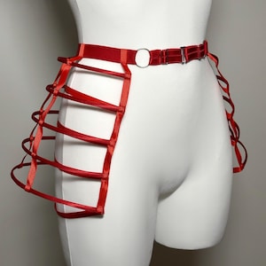 May include: A red waist harness with a cage-like design. The harness features a belt with a silver-colored buckle and a circular ring. Vertical red straps create a cage effect on the sides. The harness is displayed on a white mannequin.