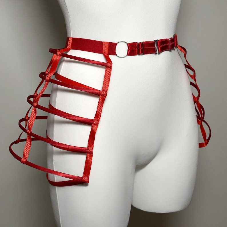 May include: A red waist harness with a cage-like design. The harness features a belt with a silver-colored buckle and a circular ring. Vertical red straps create a cage effect on the sides. The harness is displayed on a white mannequin.