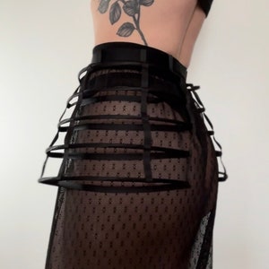 May include: Black cage skirt with a sheer, dotted fabric. The skirt features a structured cage design with horizontal bands and vertical supports, creating a unique silhouette. The skirt is made of a sheer, dotted fabric.