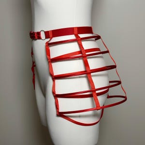 May include: Red harness-style body accessory. The piece features a red belt with a metal ring and a cage-like design made of red ribbons. The accessory is displayed on a white mannequin.