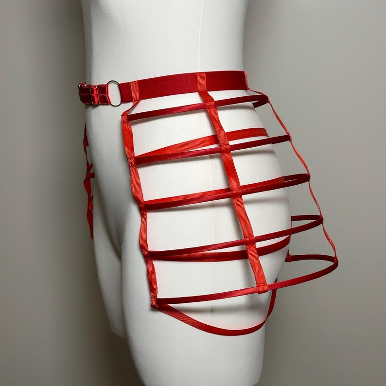 May include: Red harness-style body accessory. The piece features a red belt with a metal ring and a cage-like design made of red ribbons. The accessory is displayed on a white mannequin.