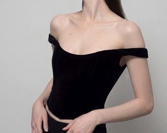 May include: A black off-the-shoulder top made of a velvet-like material. The top has a fitted bodice and a curved neckline. The sleeves are short and extend slightly over the shoulders. The top is paired with black bottoms.