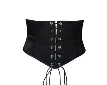 May include: Black corset with a lace-up front. The corset features silver grommets and a tie closure. The garment is made of a smooth, matte fabric and has a structured design. The corset is designed to cinch the waist.