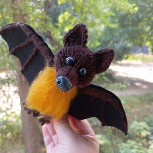 Puede incluir: Un murciélago de punto marrón con grandes ojos negros y un pecho de piel amarilla. El murciélago tiene alas de fieltro negro.
